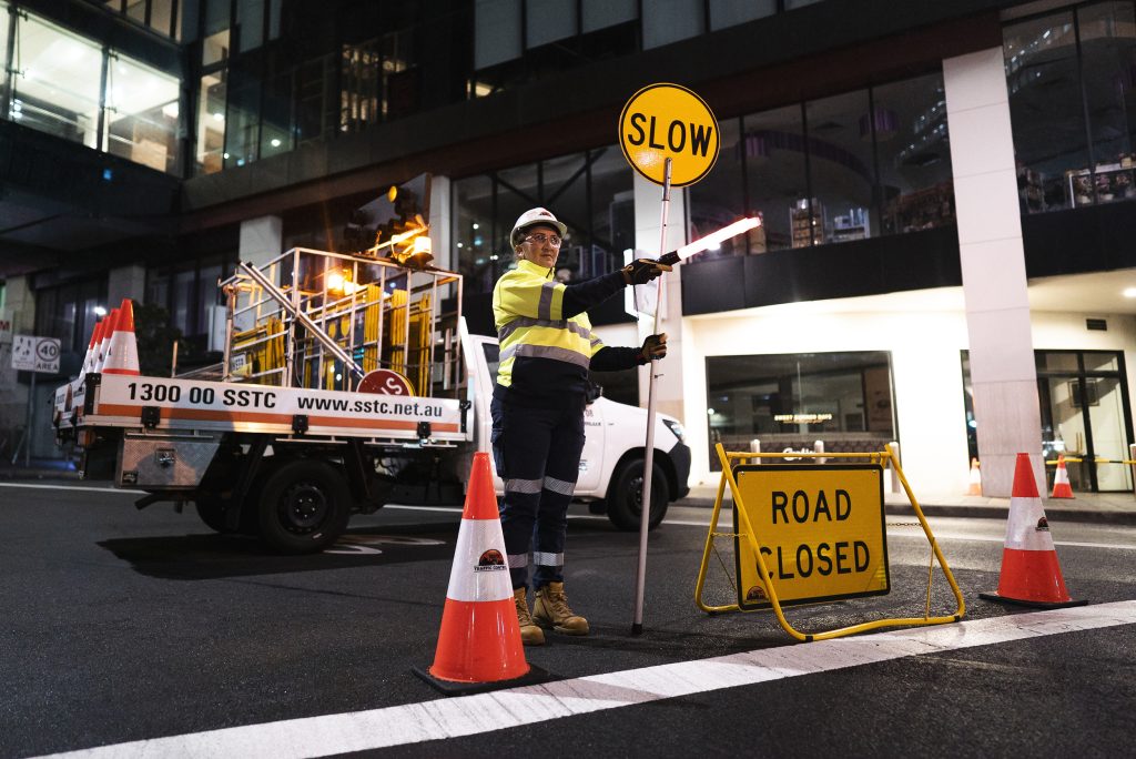 An SSTC traffic controller holding a slow sign with other traffic control equipment around her. There is a SSTC Ute in the background.