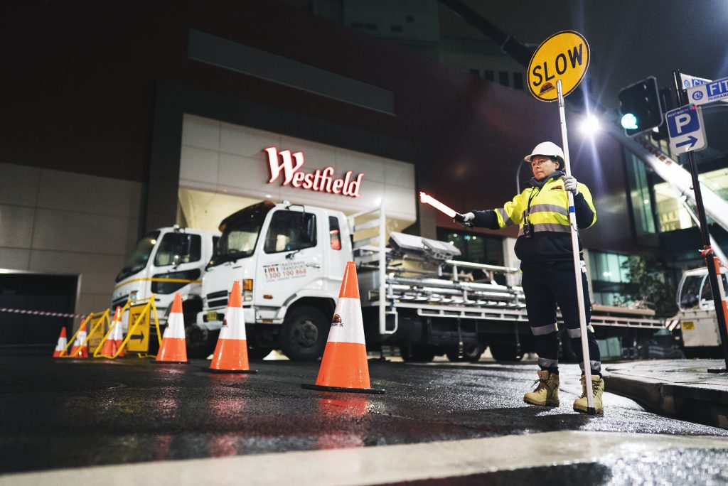 An SSTC traffic controller standing outside a Westfield shopping centre directing traffic with a slow sign. The road shows signs of rain and storms. 
