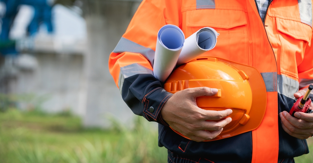 A construction worker holding a hardhat, radio and building plans under his arm. He is wearing Hi-Vis.